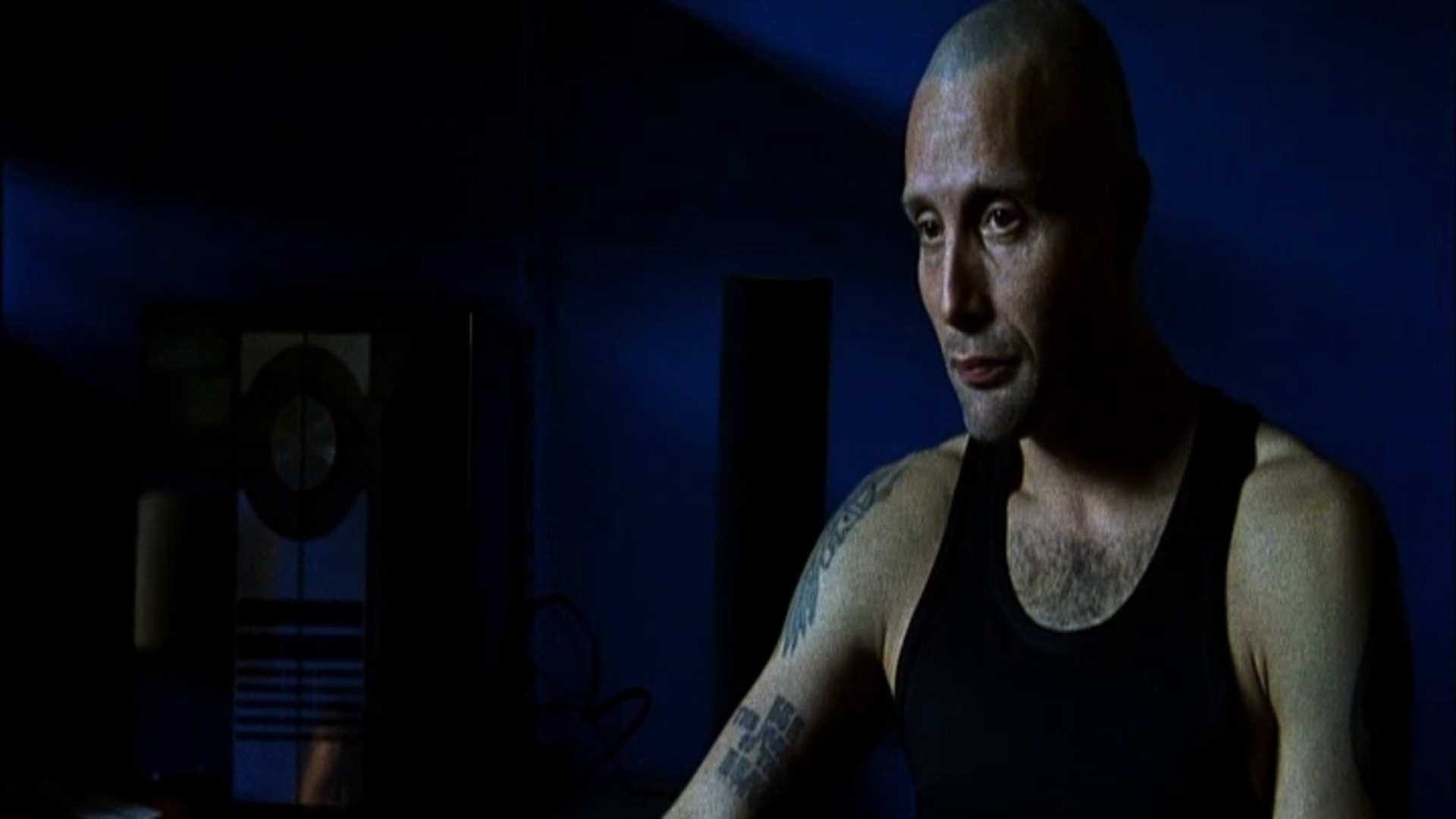 Mads Mikkelsen as Tonny seated in blue light with visible shoulder and arm tattoos