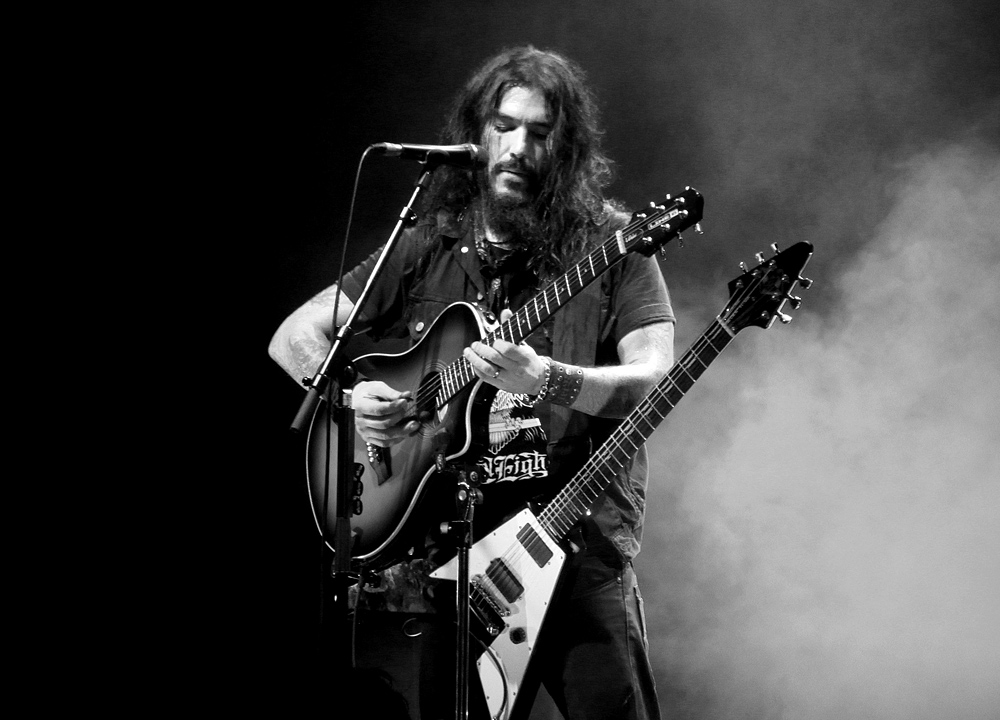Robb Flynn tattoos: clear concert photograph showing the large upper-arm panel, lower-arm imagery and wrist area without a generic portrait crop