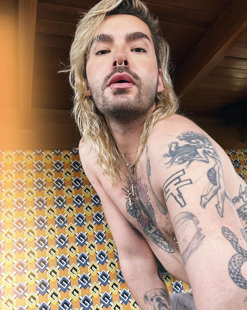 Bill Kaulitz tattoos: sharp shirtless selfie showing the central chest emblem, anatomical heart edge, bold upper-arm figures and lower arm ink