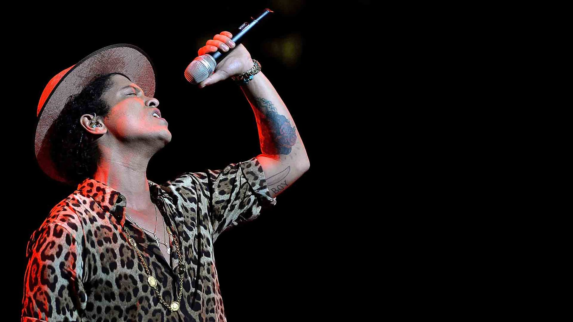 Bruno Mars tattoos: high-resolution stage image showing the layered traditional tattoo coverage across Bruno Mars’s raised forearm