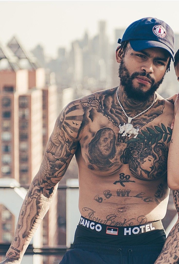 Dave East tattoos: high-resolution shirtless rooftop portrait showing chest portraits, stomach lettering and arm coverage