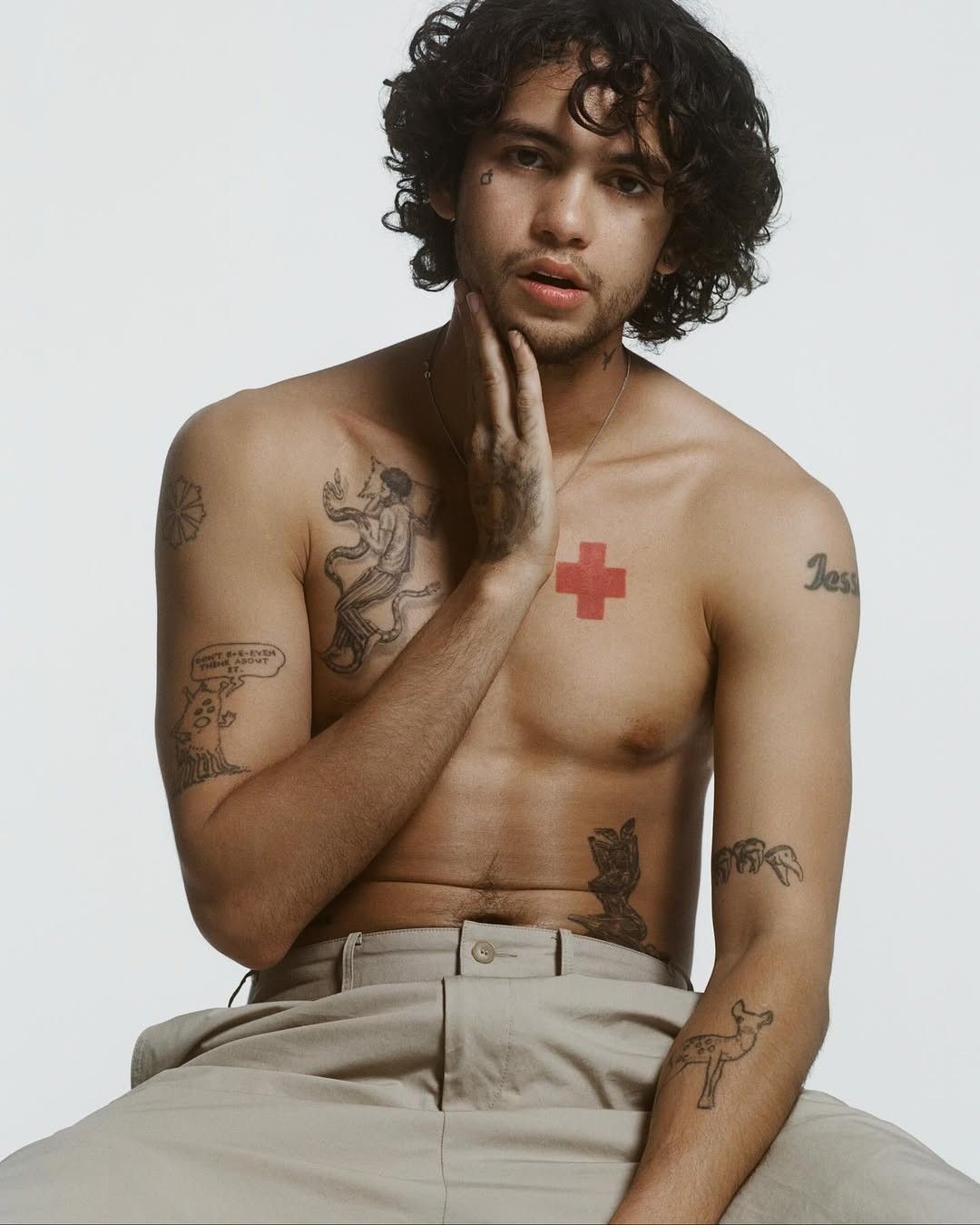 Dominic Fike tattoos: red cross, chest figure, shoulder lettering, and arm artwork