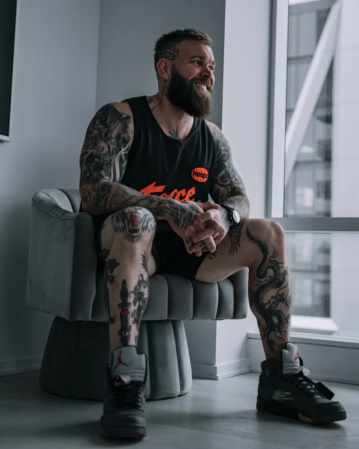 Jordan Shallow tattoos: Inked seated portrait showing Jordan Shallow’s face-visible full collection across the shoulders, arms, hands and legs
