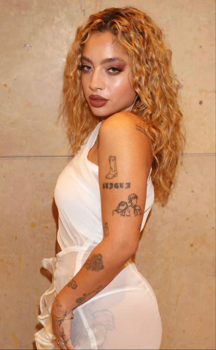 Kiana Ledé tattoos: Arm portrait showing boot, lettering and small figure tattoos; not used to identify the matching blueberry or heart designs