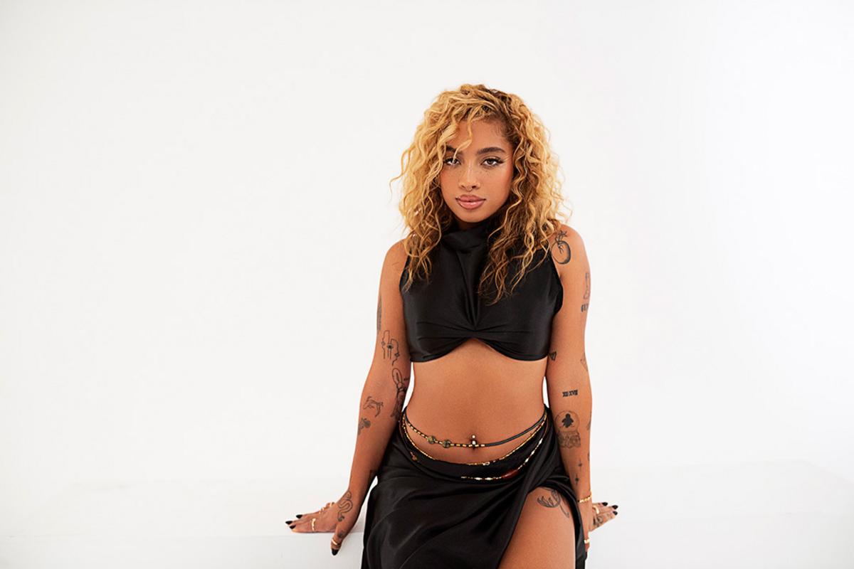 Kiana Ledé tattoos: Seated studio photo showing arm and leg tattoo coverage