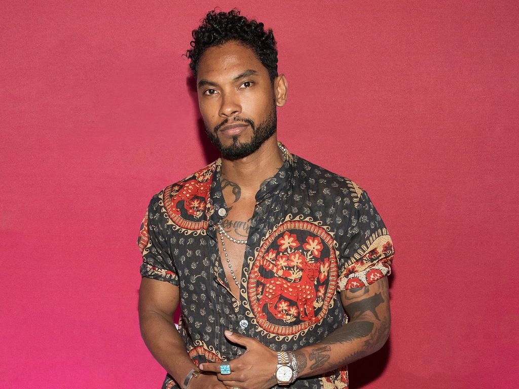 Miguel tattoos: chest lettering, neck ink and visible arm tattoos in a red studio portrait