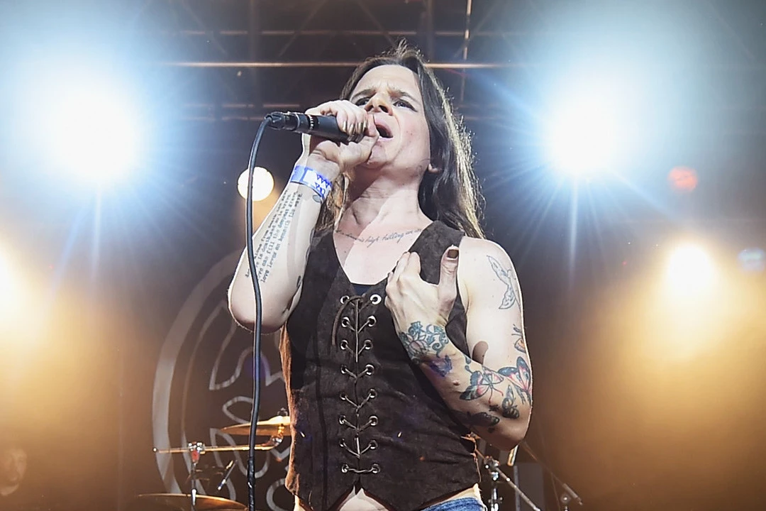 Mina Caputo tattoos: Loudwire performance photograph clearly showing the coloured hand and wrist work, chest script, upper-arm figure and surrounding tattoos.