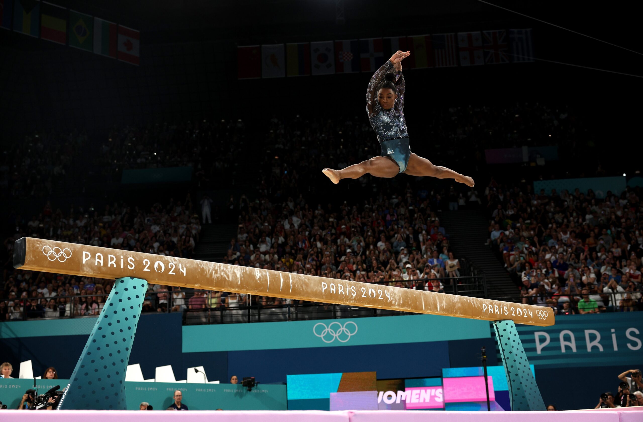 Simone Biles contextual balance-beam split leap; does not show tattoo evidence Simone Biles in a contextual photograph: contextual balance-beam split leap; does not show tattoo evidence; not tattoo evidence