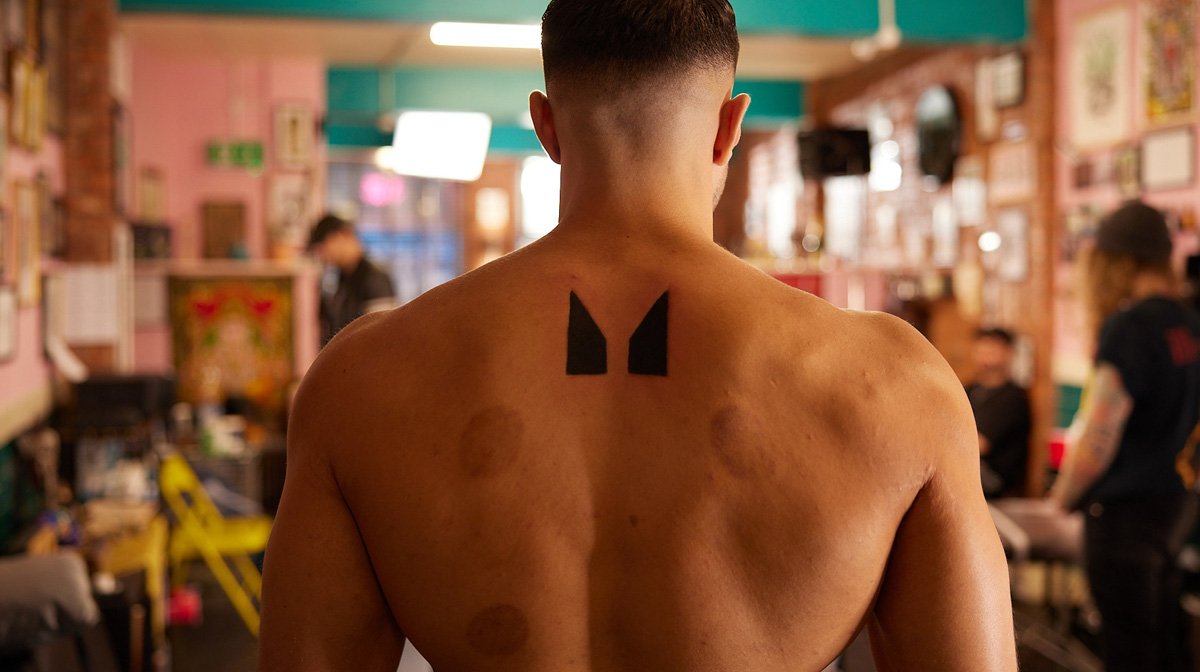 Tommy Fury tattoos: Tommy Fury photographed from behind in the tattoo studio; two solid black pointed shapes are clearly visible high on the upper back.